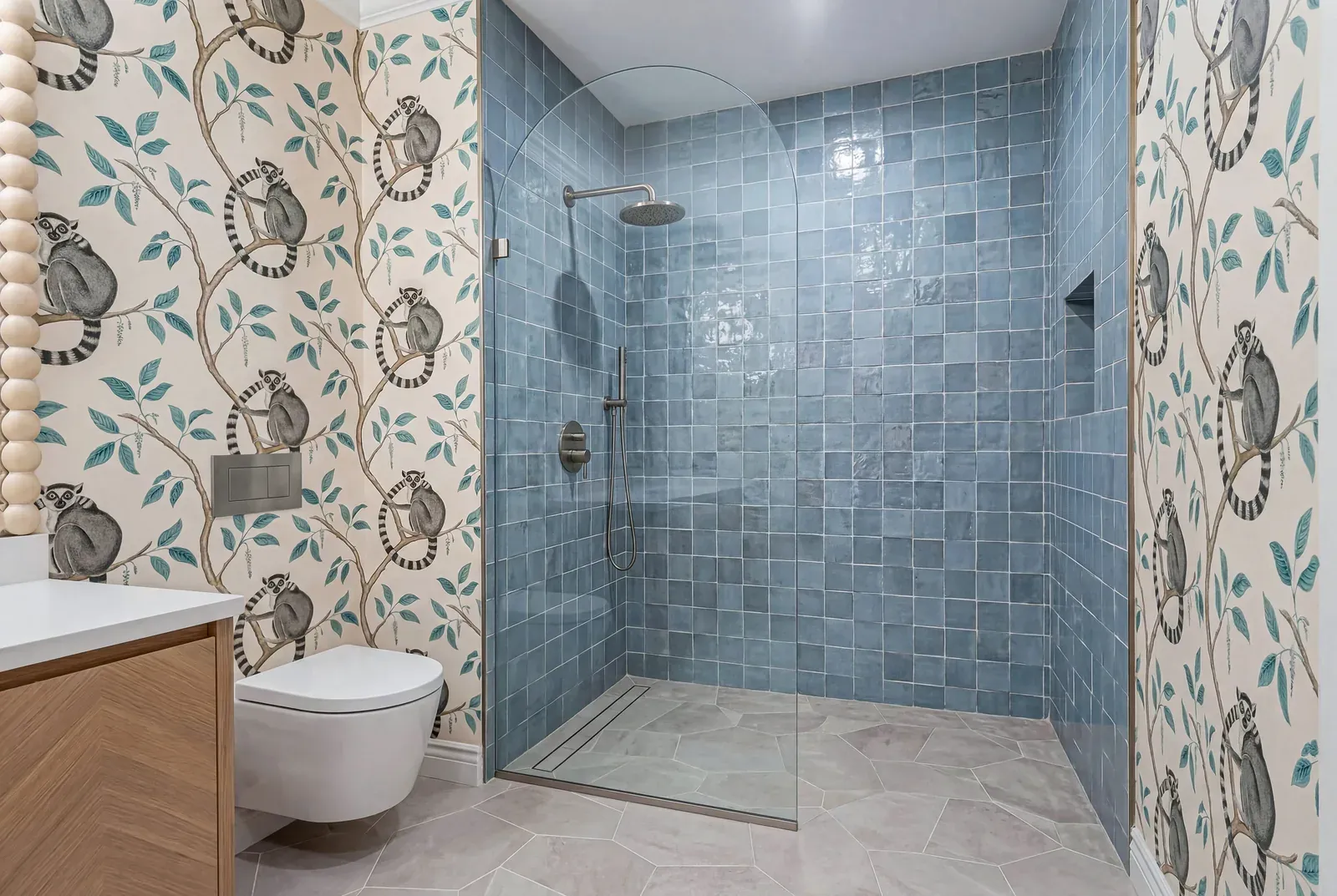 Walk-in shower tiled in blue zellige beside lemur-patterned wallpaper, a wall-hung toilet and an oak vanity