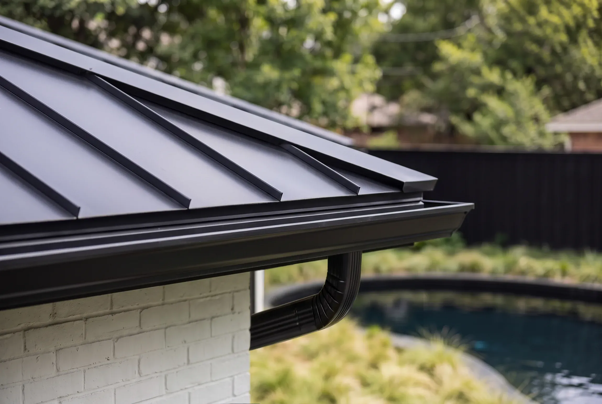Close detail of a black standing-seam metal roof edge, gutter, and downspout above a painted brick wall