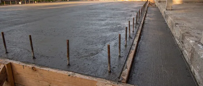 Freshly poured concrete slab with rebar dowels set along the wood form board, beside an older concrete wall