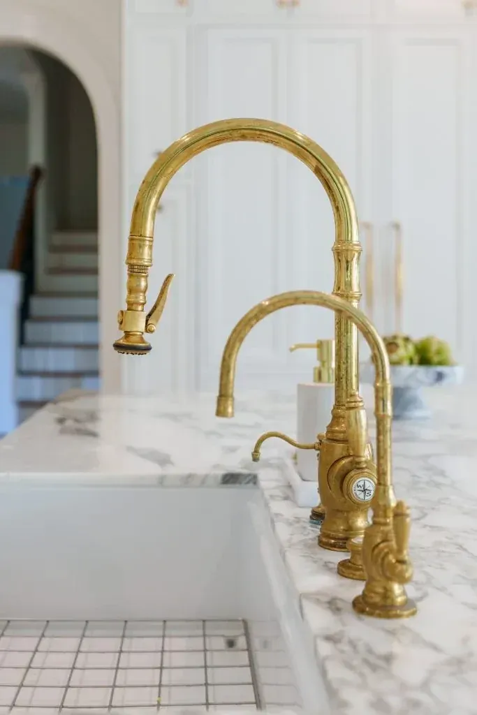 Unlacquered brass faucet set over a farmhouse sink with marble countertop