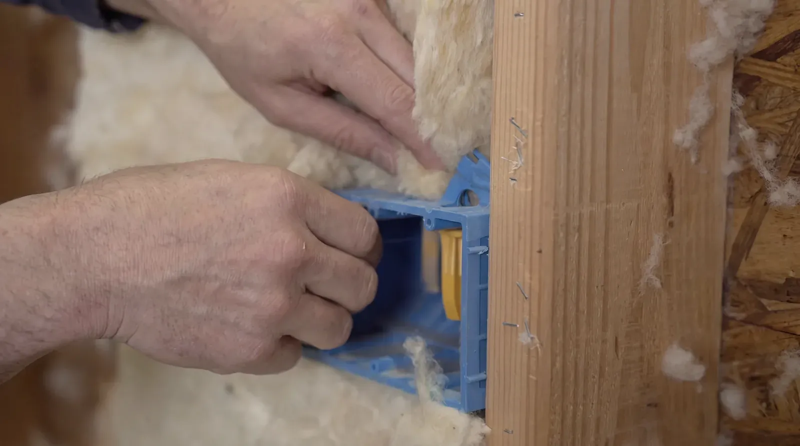 Hands tucking Havelock sheep's wool fibers around an electrical outlet box between timber studs